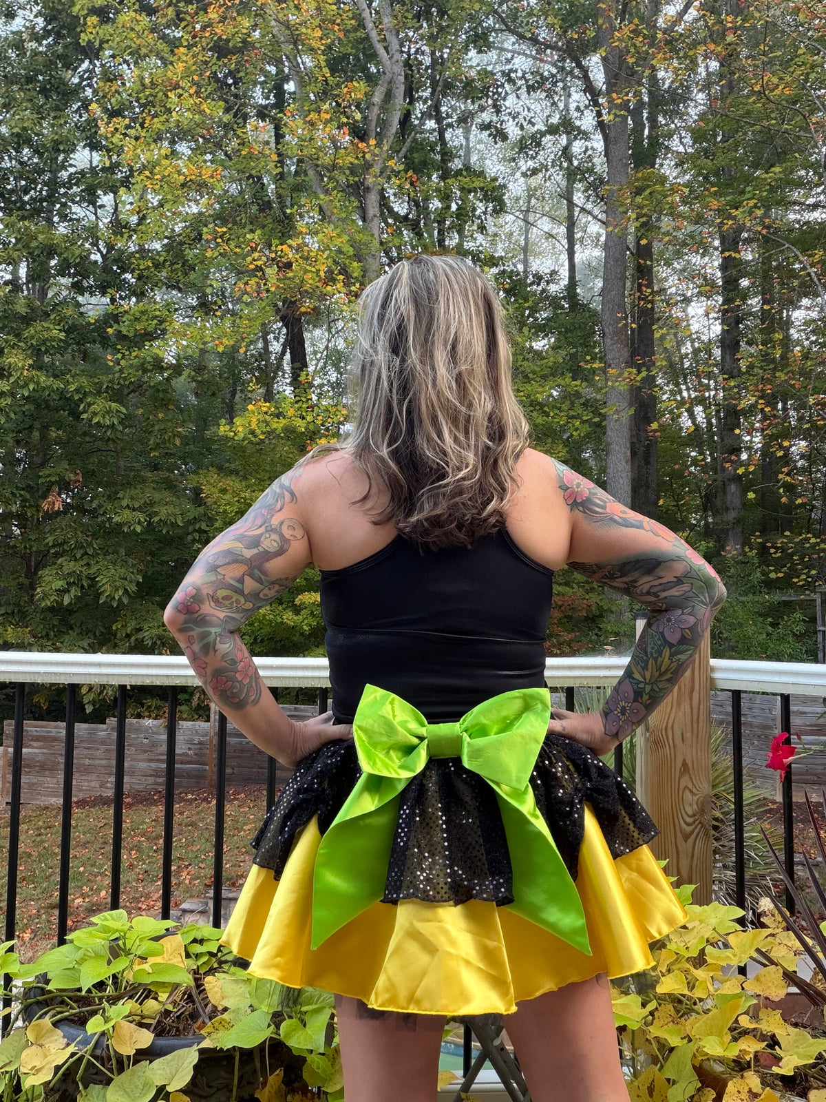 A woman stands with her back to the camera on a wooden deck, wearing a black sleeveless top and a tiered yellow and black skirt with a large lime green bow.