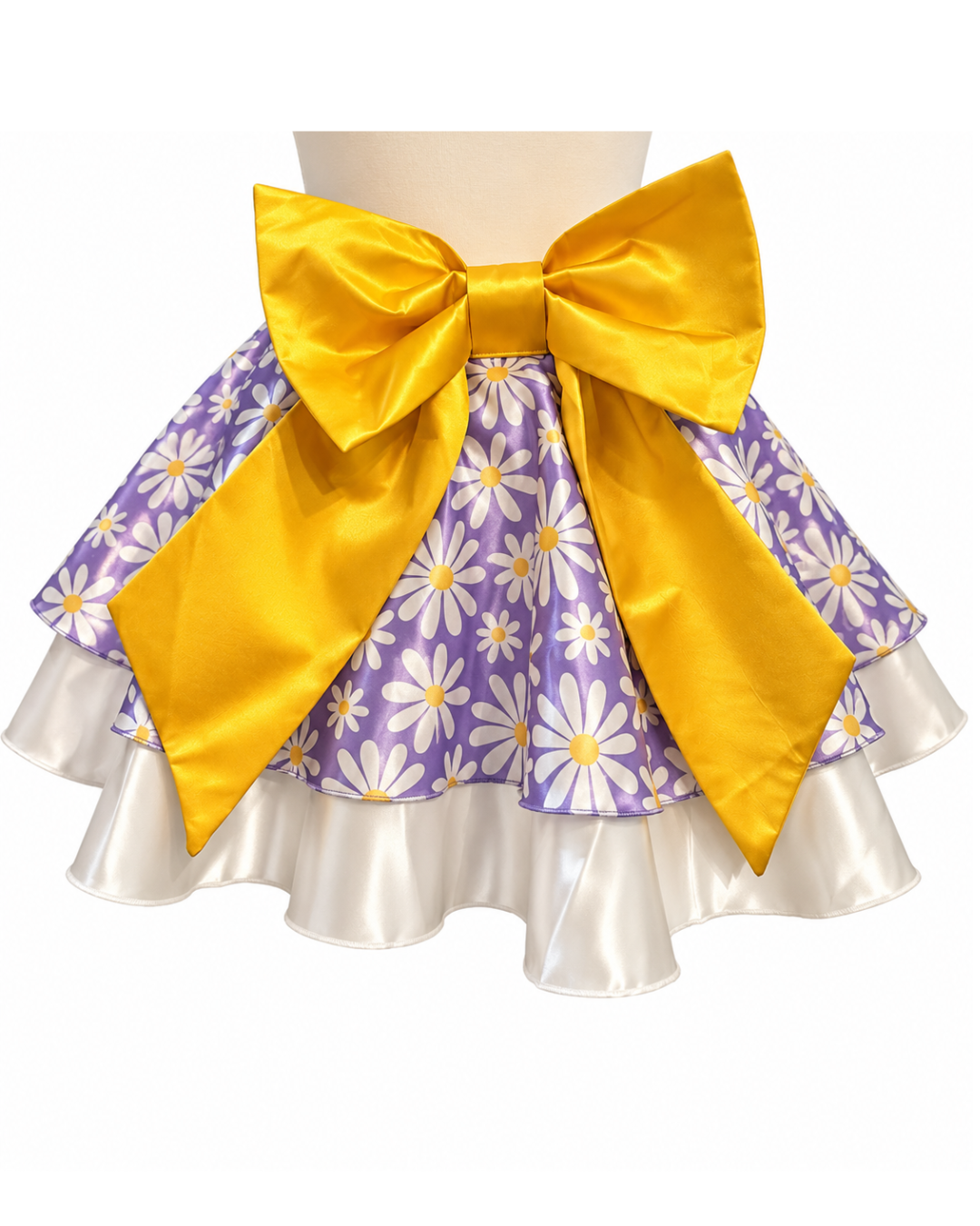 A tiered ruffle skirt with a purple and white daisy pattern, featuring a large shiny gold bow at the waistband and a white satin ruffled underskirt.