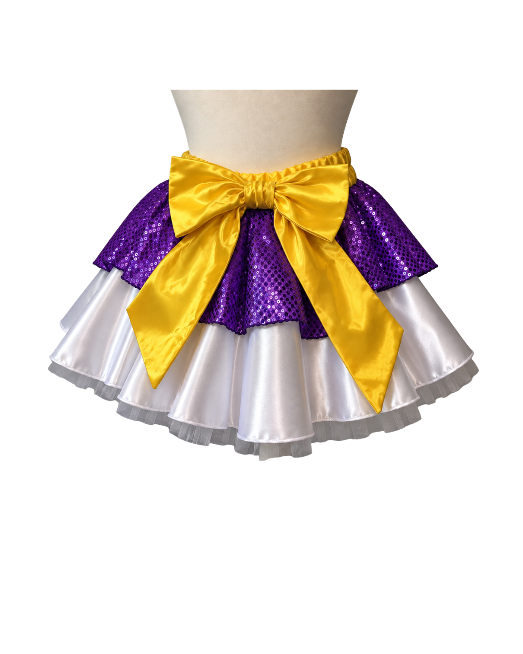 A three-layered tutu skirt with a purple sequined middle layer, a white satin bottom layer, and a large yellow satin bow at the waist, displayed on a mannequin torso against a white background.