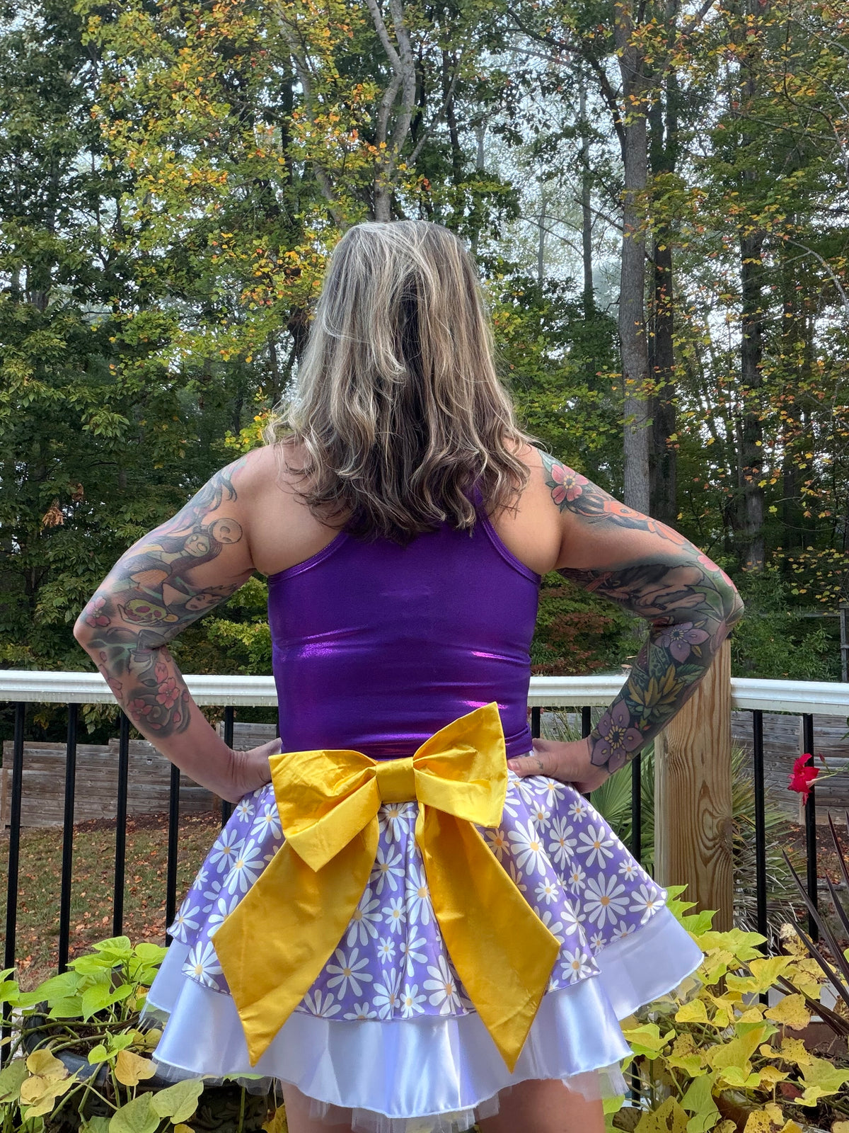 A person standing outdoors on a deck, photographed from the back, wearing a shiny purple sleeveless top and a white ruffled skirt with a large yellow bow and yellow side panels.