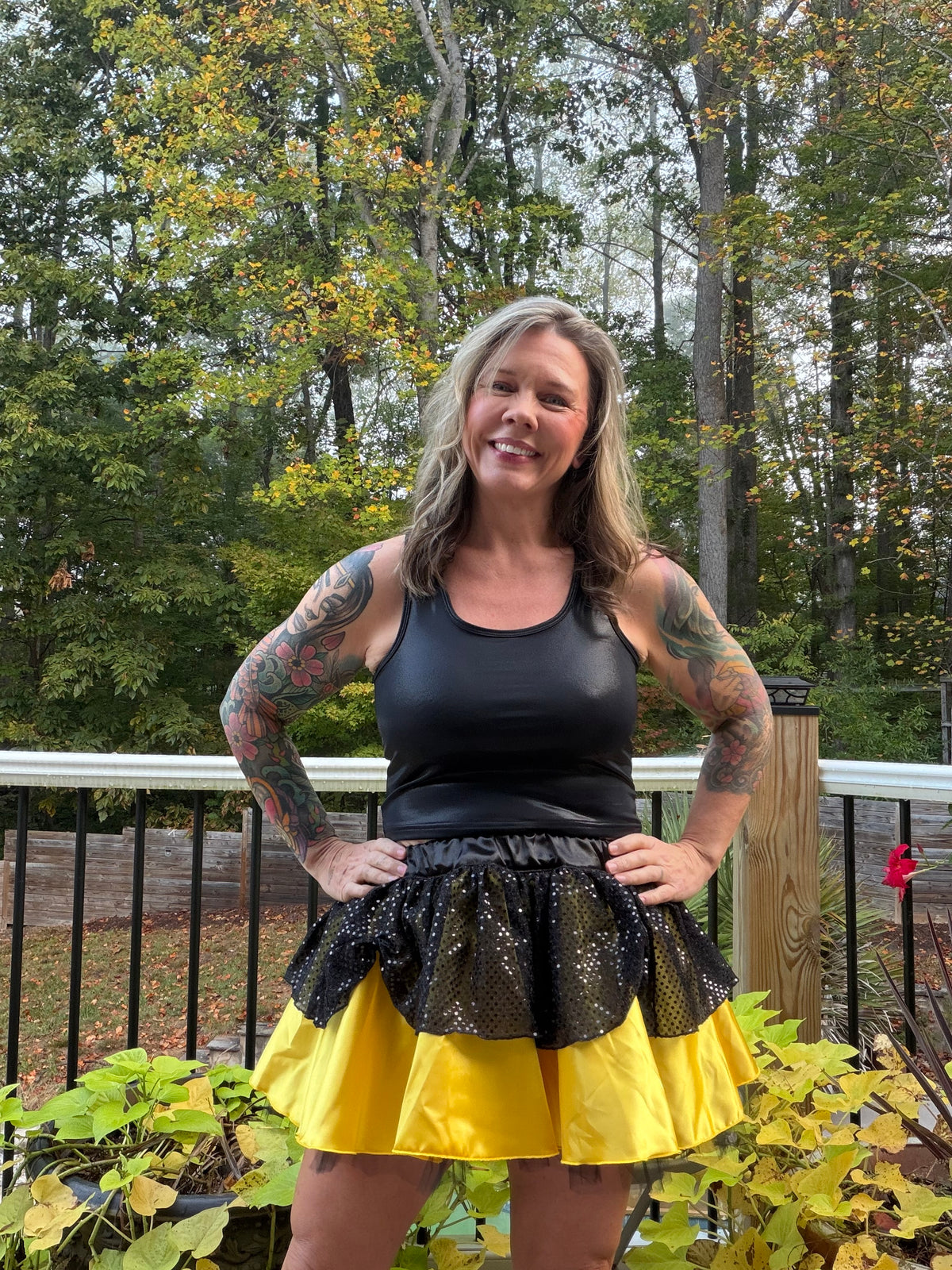 A woman with tattoos stands on a wooden deck outdoors, wearing a black sleeveless top and a black-and-yellow skirt with sequined ruffles. Trees and foliage are visible in the background.